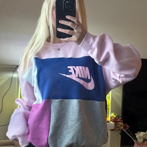 May include: A color-blocked sweatshirt featuring the Nike logo in pink. The sweatshirt has panels of pink, blue, light blue, purple, and gray. The sleeves are a solid pink color.