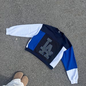 May include: Color-block sweatshirt with the letters "LA" in gray. The sweatshirt features white, blue, and black panels. The sleeves are white and blue. The garment is lying on a gray surface.