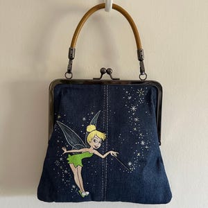May include: A denim handbag with a wooden handle and a metal clasp. The bag features an embroidered image of a fairy with a wand, surrounded by stars. The bag is approximately 8 inches wide.