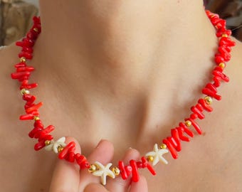 Peut inclure: Un collier en corail rouge orné de perles blanches en forme d'étoiles de mer et de petites perles dorées. Le collier est tenu par une personne portant un débardeur blanc. Un accessoire vibrant.