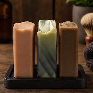 May include: Three rectangular bars of soap standing upright on a black tray. The left bar is solid peach, the center bar features a marbled pattern of white, sage green, and dark gray, and the right bar is a solid, textured earthy brown color.
