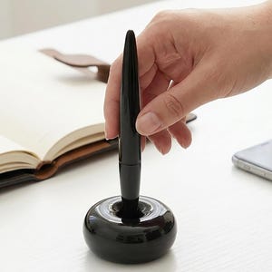 May include: A black pen in a matching pen holder. The pen is held upright in a round, glossy black base. A hand is holding the pen. A leather-bound notebook and a smartphone are in the background.