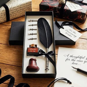 May include: A calligraphy set in a black box, featuring a black feather quill, ink bottle, inkwell, and several nibs. The ink bottle reads "Calligraphy Set - Ink". Gift boxes and tags with the text "For the Discerning Writer" are also present.