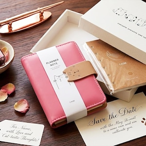 May include: A pink planner with a cat design, a brown leather strap, and the words "PLANNER NOTE" on a white band. The planner is in a white box with a cat illustration. A card reads "Save the Date".