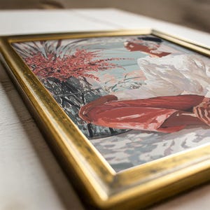 May include: Framed artwork featuring a seated figure in white and rust-colored clothing. The painting has a gold frame and depicts a person in a serene outdoor setting with plants and a light blue background. The artwork is a painting.