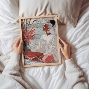 May include: A framed art print featuring a person in a white sweater and red pants, seated in a meditative pose. The artwork has a light wood frame and depicts a serene scene with plants and a window view. The person is wearing a headscarf.