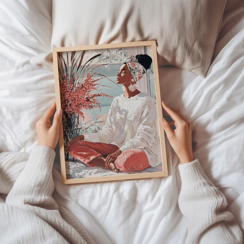 May include: A framed art print featuring a person in a white sweater and red pants, seated in a meditative pose. The artwork has a light wood frame and depicts a serene scene with plants and a window view. The person is wearing a headscarf.