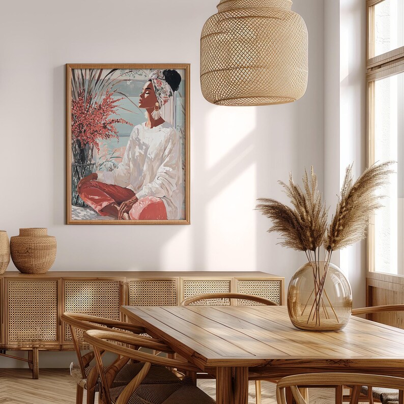 May include: A framed art print depicts a person in a white top and red pants, seated in a meditative pose. The artwork is displayed in a room with a wooden table, chairs, and a woven light fixture. A vase of dried plants sits on the table.