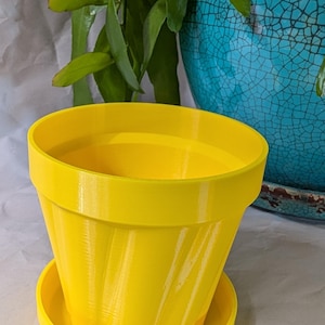 Puede incluir: Una maceta amarilla brillante con un platillo a juego. La maceta tiene un diseño acanalado y está vacía. Una maceta de cerámica azul y plantas verdes están en el fondo.
