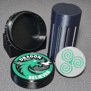 May include: An assortment of 3D-printed items, including a black container with a lid, a dark blue cylindrical container, and two circular objects. One circular object features a green dragon design with the text "DRAGON BELIEVER." The other has a green spiral design.