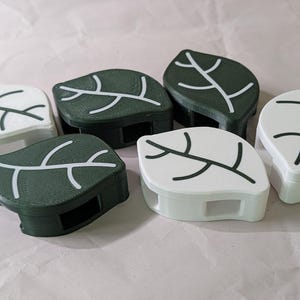 May include: Six leaf-shaped decorative boxes. Three are white with black and white leaf vein designs, and three are dark green with white leaf vein designs. The boxes have a rectangular base with a cutout.