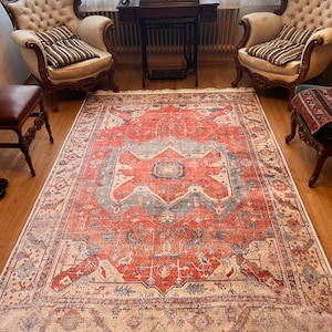 Pode incluir: Um tapete vermelho, azul e creme com um design persa tradicional. O tapete tem uma borda com franjas e está colocado sobre um piso de madeira.