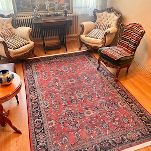 Puede incluir: Una alfombra persa roja, azul y crema con un patrón floral. La alfombra está colocada sobre un suelo de madera en un salón con dos sillas antiguas y una mesa.