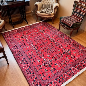 May include: A red and black patterned rug with a white fringe. The rug is laid out on a wooden floor in a room with two antique chairs.