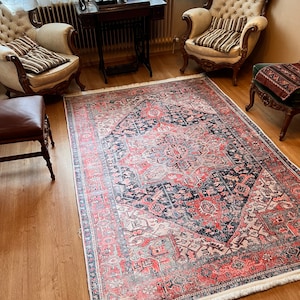 May include: A vintage-style rug with a geometric pattern in shades of pink, red, black, and white. The rug has a fringe border and is laid out on a hardwood floor in a living room setting.