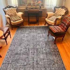 May include: A blue and gray patterned rug with a white fringe border. The rug is in a living room setting with two antique chairs and a table with a painting above it.