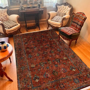 Puede incluir: Una alfombra antigua, rectangular y grande, con un campo predominantemente marrón rojizo. La alfombra presenta un detallado diseño floral en tonos azules, verdes y crema, con un borde azul contrastante. La alfombra está colocada sobre un suelo de madera.