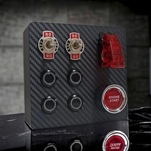 May include: A black panel with a carbon fiber pattern, featuring four round toggle switches, two red toggle switches labeled "NO", and a red push button labeled "ENGINE START".