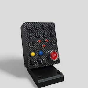 May include: A black control panel with a carbon fiber texture, featuring numerous black buttons, red, yellow, and blue buttons, and a red "ENGINE START" button. The panel is mounted on a black base.