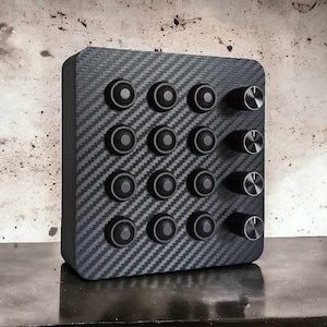 May include: A rectangular black control panel with a carbon fiber pattern. It features 12 round black buttons and 4 silver knobs. The panel is designed for electronic devices and has a modern, industrial aesthetic.