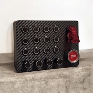 May include: A black control panel with a carbon fiber pattern. It features multiple black buttons, knobs, and a red "ENGINE START" button. A red toggle switch is also visible.
