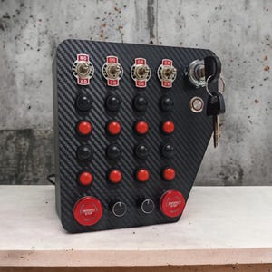 May include: A black panel with a carbon fiber pattern, featuring multiple red and black push buttons, four toggle switches labeled "ON", and two red emergency stop buttons. The panel also includes two knobs and a keyhole with a key attached.