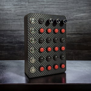 May include: A black box with a carbon fiber pattern and multiple buttons and knobs. The buttons are red and black, and the knobs are black.