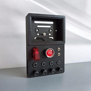 May include: A black control panel with a carbon fiber texture. It features a red "ENGINE START" button, toggle switches, and rotary knobs. A red, rectangular switch is also present. The panel has an open top section with mounting holes.