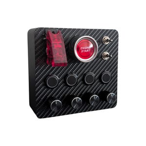 May include: A rectangular control panel with a carbon fiber pattern finish. It features a red engine start button, a red flip-up safety cover, two metal toggle switches, four black circular push buttons, and four black rotary control knobs.