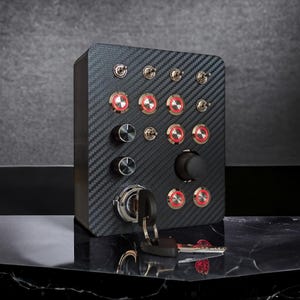 May include: A black panel with a carbon fibre texture features multiple red and black toggle switches, a black knob, and a keyhole. The panel is mounted on a black surface.