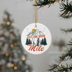 May include: White ceramic Christmas ornament with a mountain and forest design. The ornament features the text "JUST ONE MORE Mile" in red and white. A gold string is attached for hanging on a Christmas tree.