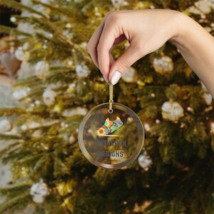 Puede incluir: Un adorno navideño redondo y transparente con un diseño de zapato colorido y el texto "UNT HERLIN DESIGNS". El adorno es sostenido por una mano frente a un árbol de Navidad.