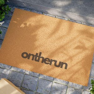 以下が含まれることがあります： 黒い文字で「ontherun」と書かれた茶色のココナッツ製のドアマットです。
