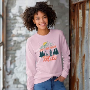 May include: A light pink long-sleeve shirt featuring a graphic print of a mountain range, a yellow sun, and green pine trees. The text "JUST ONE MORE" appears in orange block letters above the word "Mile" in a cursive orange script font.