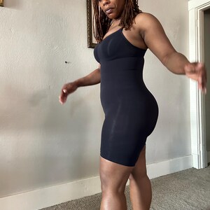 May include: A black, one-piece bodysuit with a built-in bra and shorts. The garment has thin straps and a form-fitting design. The fabric appears smooth and stretchy. The bodysuit is designed to be worn as shapewear or as a base layer.