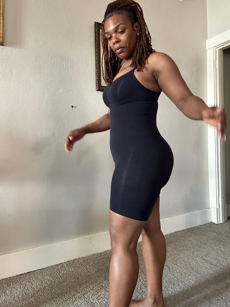 May include: A black, one-piece bodysuit with a built-in bra and shorts. The garment has thin straps and a form-fitting design. The fabric appears smooth and stretchy. The bodysuit is designed to be worn as shapewear or as a base layer.