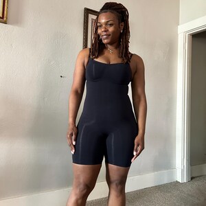 May include: A black bodysuit with spaghetti straps and built-in bra cups. The shorts-style bottom extends to mid-thigh. The garment is made of a smooth, form-fitting material. The model is standing in a room with neutral-colored walls and carpet.