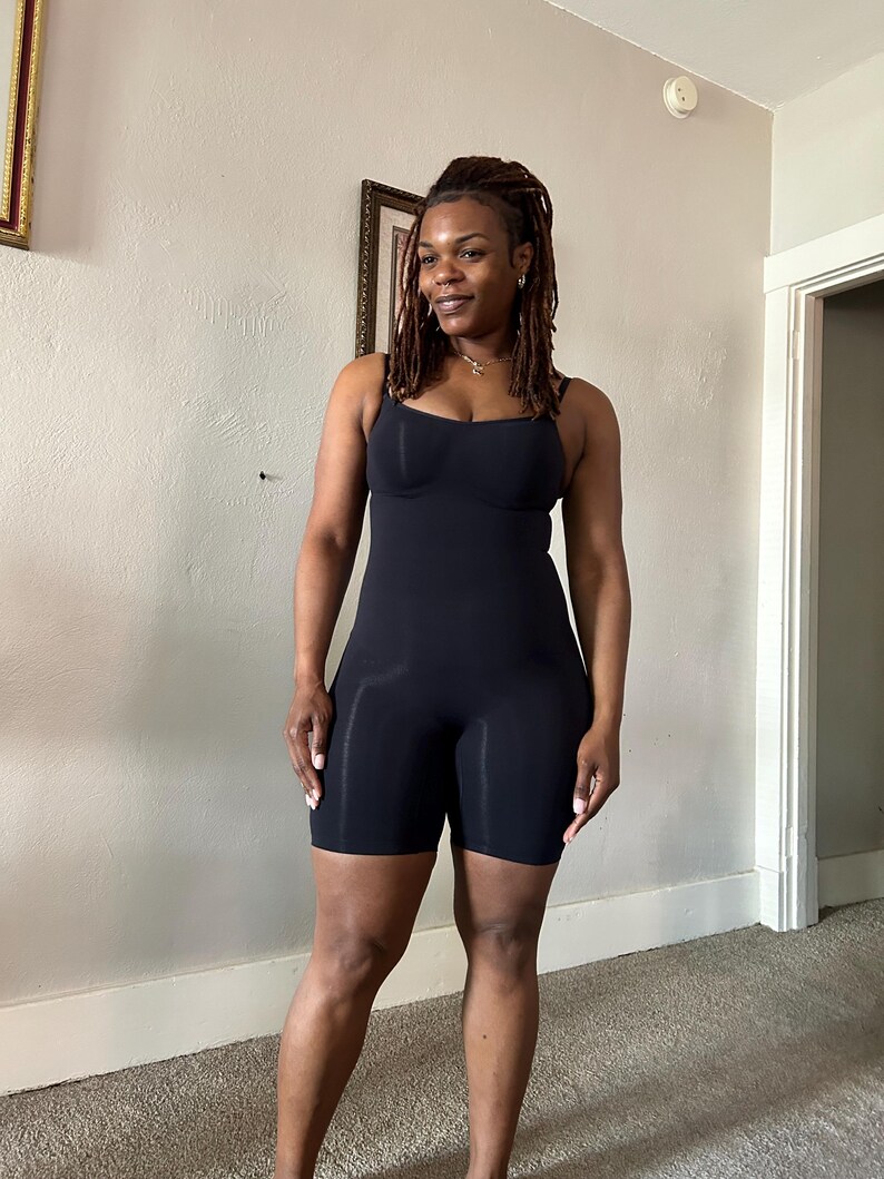 May include: A black bodysuit with spaghetti straps and built-in bra cups. The shorts-style bottom extends to mid-thigh. The garment is made of a smooth, form-fitting material. The model is standing in a room with neutral-colored walls and carpet.
