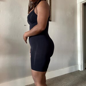 May include: A black, sleeveless bodysuit with a scoop neckline and built-in shaping. The garment extends to mid-thigh, offering a smooth silhouette. The fabric appears to be a stretchy, form-fitting material.