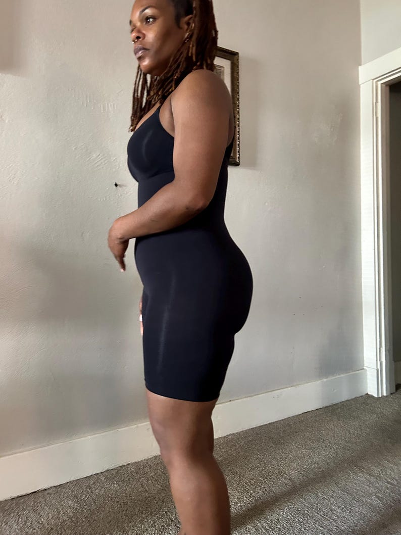 May include: A black, sleeveless bodysuit with a scoop neckline and built-in shaping. The garment extends to mid-thigh, offering a smooth silhouette. The fabric appears to be a stretchy, form-fitting material.