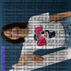 May include: White t-shirt featuring a cartoon illustration of a girl in a baseball cap and uniform. The girl has pink hair and is depicted in a playful pose. The design is printed on a white, short-sleeved t-shirt. The t-shirt is shown on a model, partially visible. The background is a blurred, dark blue color with repeating text. The overall style is casual and playful, suitable for everyday wear.