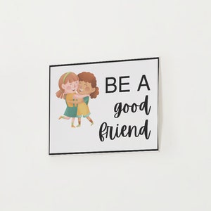 May include: A square art print with a black border, featuring the text "BE A good friend" in a handwritten style. The print includes an illustration of two girls hugging, with one wearing a green dress and the other a yellow dress.