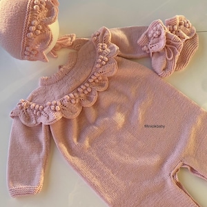 May include: A pink knitted baby outfit with a ruffled neckline and matching hat and booties. The outfit is decorated with small white pom-poms.