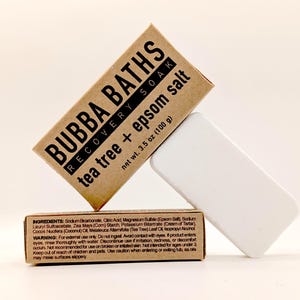 May include: A product shot features two rectangular boxes and a white bar. The boxes are brown with black text that reads "BUBBA BATHS" and "tea tree + epsom salt." The white bar is a solid, rectangular shape. The net weight is 3.5 oz (100 g).