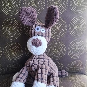 May include: A brown crocheted dog toy with white accents. The dog has large, white eyes and a brown nose. The toy is sitting on a brown patterned surface.