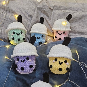 May include: Six crocheted boba tea plushies in various colors, including purple, yellow, pink, blue, and green. Each plush has a white crocheted lid and black crocheted straw. The plushies are arranged on a blue and gray fabric background with string lights.