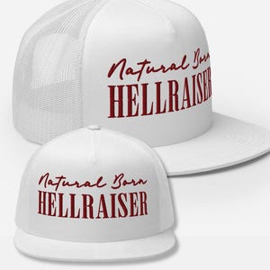 May include: White trucker hat with a mesh back and a flat brim. The hat features the text "Natural Born HELLRAISER" in a bold, burgundy script across the front. A second hat is in the foreground.