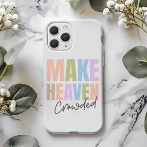 Puede incluir: Funda de teléfono blanca con el texto "MAKE HEAVEN Crowded" en colores pastel. La funda está rodeada de flores blancas y hojas verdes sobre una superficie de mármol.