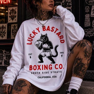 May include: White long-sleeve shirt featuring a boxing cat graphic and the text "LUCKY BASTARD BOXING CO." The shirt also has the text "SOUTH SIDE STRAYS CALIFORNIA, USA". The shirt is worn by a person with tattoos.