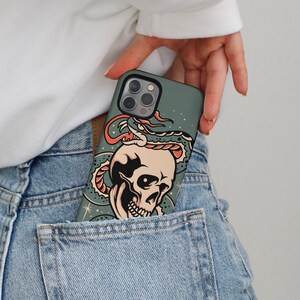 Puede incluir: Funda de teléfono verde azulado con un diseño de calavera y serpiente. La calavera es beige con detalles negros, y la serpiente es roja y verde azulado. La funda está en un bolsillo vaquero.